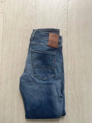 Blå Replay jeans  - Säljer ett par blå jeans från Replay med klassisk femficksmodell och snygg skinnpatch bak i midjan. Jeansen har en smal passform och diskreta slitningar för en avslappnad look. Materialet är mjukt denim med stretch för extra komfort.