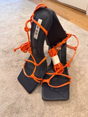 Svarta pumps med orange snörning NA-KD - Säljer ett par trendiga svarta pumps från NA-KD med fyrkantig tå och höga klackar. Skorna har långa orange snörningar som kan lindas runt benet för en cool look. Tillverkade i syntetmaterial och har en unik, modern design.