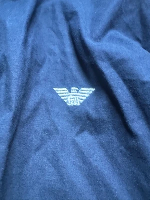 Mörkblå skjorta från Armani - Snygg mörkblå skjorta från Armani med klassisk knappstängning och det ikoniska Armani-logotypen broderad på bröstet. Skjortan är tillverkad i ett mjukt bomullstyg och har en stilren design som passar perfekt till många olika looks. Knapp vid vänster arm är borta.