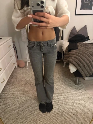 Grå bootcut jeans med låg midja - Säljer ett par gråa bootcut jeans med låg midja och dubbla knappar fram. Jeansen har klassisk femficksdesign och är tillverkade i ett mjukt denimtyg. Jeansen har däremot en uppklippt sömn på vardera ben längst ner men är annars i ett bra skick.