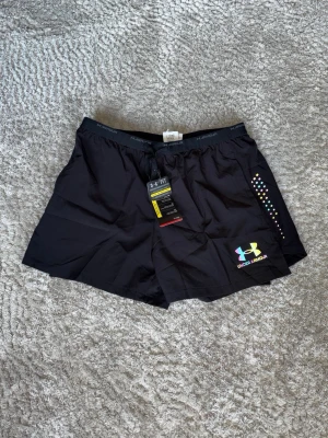 Svarta träningsshorts från Under Armour - Svarta träningsshorts från Under Armour med elastisk midja och färgglad logga framtill. Shortsen har diskreta färgprickar på sidan och är tillverkade i ett lätt och snabbtorkande material som passar perfekt för träning.