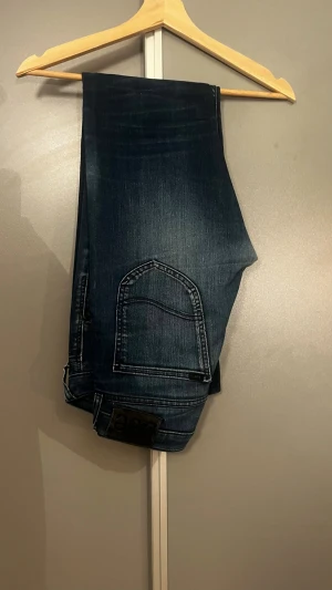 Mörkblå slim fit jeans från Lee - Snygga mörkblå jeans från Lee med slim fit passform och klassiska fem fickor. Jeansen har diskreta slitningar och kontrastsömmar som ger en cool look. Tillverkade i ett mjukt denimtyg som sitter skönt hela dagen.