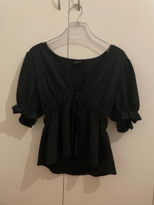 Svart blus med snörning från Gina Tricot - Säljer en svart blus från Gina Tricot med v-ringning, puffiga korta ärmar och snörning framtill. Blusen har volangdetaljer vid ärmslut och midja, vilket ger en feminin och trendig look. Perfekt att styla med jeans eller kjol.