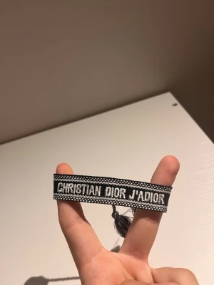 Christian Dior J'Adior armband - Snyggt svart och vitt tygarmband från Christian Dior med texten 'J'ADIOR' broderad framtill. Armbandet har justerbara band med tofsar i ändarna och en cool, trendig look som passar perfekt till din outfit.