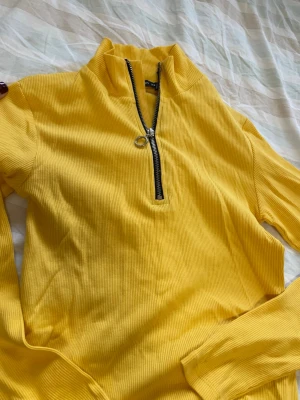 Gul ribbad tröja med half zip - Gul långärmad tröja från FB Sister i ribbad bomull med en snygg half zip-dragkedja och hög krage. Perfekt för dig som gillar färg och vill ha en bekväm men trendig look. Passar till jeans eller kjol för en cool vardagsstil.