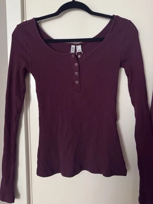 Vinröd topp - Snygg vinröd ribbad topp med lite lila ton, långärm och knappar framtill. Toppen är i ett förvånat stretchigt material och är otroligt skön mot huden. Sjukt snyggt att styla med ett vitt linne under. Helt oanvänt och prislappen kvar, i storlek S men passar även XS💕