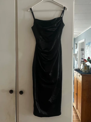 Svart långklänning med smala axelband - Snygg svart långklänning med smala axelband och draperad detalj framtill. Klänningen har en figurnära passform och är tillverkad i ett glansigt material som ger en elegant look. Perfekt för dig som vill ha en stilren och tidlös outfit.