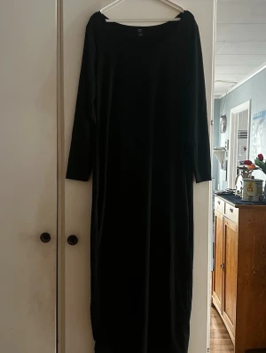 Svart långklänning med lång ärm - En enkel och stilren svart långklänning med långa ärmar och rak passform. Klänningen har en rund halsringning och är perfekt för dig som gillar minimalistisk stil. Materialet ser mjukt och följsamt ut, vilket ger en bekväm känsla.