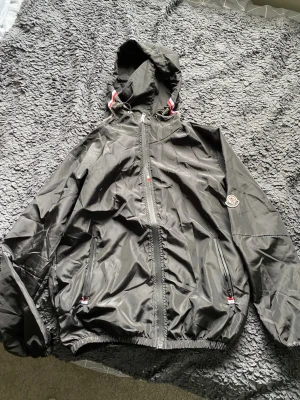 Svart vindjacka från Moncler - Snygg svart vindjacka från Moncler med huva och dragkedja framtill. Jackan har två fickor med dragkedja och elastiska muddar vid ärmslut och nederkant. Moncler-logga på vänster ärm och detaljer i rött och vitt vid huvan. Perfekt för blåsiga dagar.