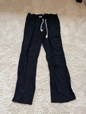 Svarta linnebyxor från Roxy - Snygga svarta linnebyxor med rak passform och vit dragsko i midjan. Byxorna har resår i midjan och är tillverkade i ett lätt och luftigt linnematerial, perfekta för en avslappnad stil. Vänstra benet har ett superliten fläck.