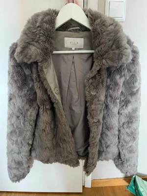 Grå fuskpälsjacka från VILA - Snygg grå fuskpälsjacka från VILA med bred krage och knäppning framtill. Jackan har ett mjukt foder och är i en kortare modell som ger en trendig look. Perfekt för att hålla värmen under kalla dagar och ger en lyxig känsla till din outfit.