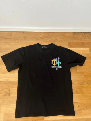 Svart Chrome Hearts t-shirt med blommor - Säljer en svart t-shirt från Chrome Hearts med färgglada blommor och logga tryckt på ryggen samt mindre tryck på bröstet. T-shirten har korta ärmar och en klassisk passform. Perfekt för dig som gillar streetwear och unika prints.