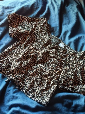Leopardmönstrad offshoulder blus - Snygg offshoulder blus med leopardmönster i beige, brunt och svart. Blusen har korta ärmar och resår vid axlarna för en trendig och bekväm passform. Tillverkad i ett lätt och luftigt tyg som ger en avslappnad känsla.