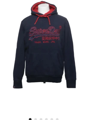 Mörkblå Superdry hoodie med röd text - Snygg mörkblå hoodie från Superdry med röd broderad logga och japanska tecken på bröstet. Hoodien har röd insida i huvan och röda dragsnören. Klassisk känguruficka framtill och ribbade muddar vid ärmslut och nederkant. Perfekt för en avslappnad streetstyle.