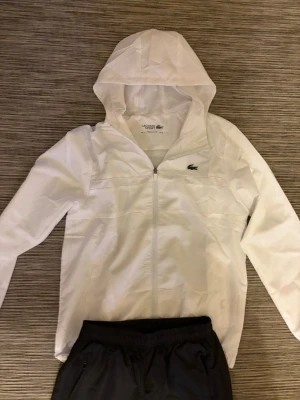 Vit vindjacka från Lacoste Sport - Snygg vit vindjacka från Lacoste Sport med huva och dragkedja framtill. Jackan har meshfoder och den klassiska gröna krokodilloggan på bröstet. Perfekt för blåsiga dagar och enkel att matcha med olika outfits.