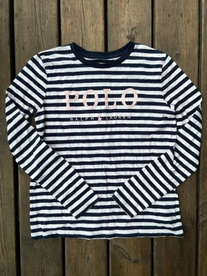 Randig långärmad topp från Polo Ralph Lauren - Snygg långärmad topp från Polo Ralph Lauren med marinblå och vita ränder. Stor logga i rosa brodyr framtill. Tillverkad i mjuk bomull, perfekt för en avslappnad och trendig look.