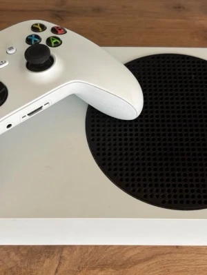 Xbox Series S spelkonsol med handkontroll - Säljer en Xbox Series S spelkonsol i vit färg med svart cirkulärt ventilationsgaller. Konsolen är kompakt och stilren, tillverkad i plast. Medföljer en matchande vit trådlös handkontroll med färgade knappar och modern design.
