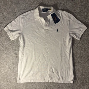Vit pikétröja från Polo Ralph Lauren - Klassisk vit pikétröja från Polo Ralph Lauren med korta ärmar och liten broderad logga på bröstet. Tröjan har krage och knappar framtill. Perfekt till en avslappnad men ändå stilren look.