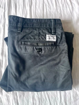 Svarta chinos från Norse Projects - Snygga svarta chinos från Norse Projects med klassisk rak passform. Byxorna har sidofickor, bakficka med knapp och är tillverkade i ett mjukt bomullstyg. Perfekta för en stilren och avslappnad look.