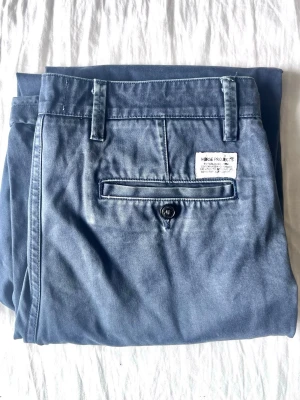 Blå chinos från Norse Projects - Snygga blå chinos från Norse Projects med klassisk rak passform. Byxorna har bälteshällor, sidofickor och en bakficka med knapp. Tillverkade i mjukt bomullstyg som ger en avslappnad men stilren look.