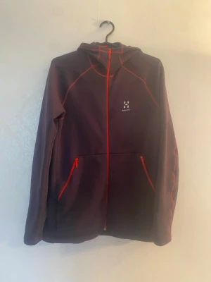 Lila hoodie från Haglöfs med dragkedja - Snygg lila hoodie från Haglöfs med röda detaljer och hel dragkedja framtill. Två fickor med röda dragkedjor och matchande röda sömmar. Tillverkad i mjukt funktionsmaterial och har en praktisk huva. Perfekt för en aktiv livsstil.