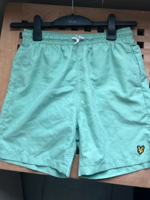 Mintgröna badbyxor från Lyle & Scott - Snygga mintgröna badbyxor från Lyle & Scott med elastisk midja och vit snörning. Klassisk gul logga på vänster ben och praktisk bakficka med lock. Perfekta för sommaren och chill dagar.