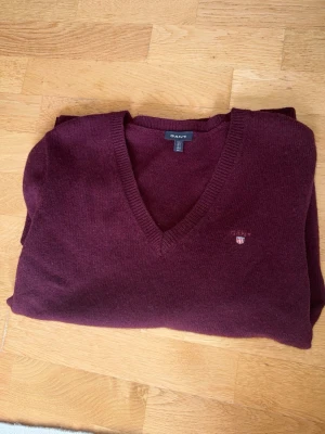 Vinröd stickad tröja från Gant - Mysig vinröd stickad tröja från Gant med V-ringning och diskret logga på bröstet. Tröjan har långa ärmar, ribbade muddar och är gjord i 100% ull. Perfekt för dig som gillar klassisk och stilren design.