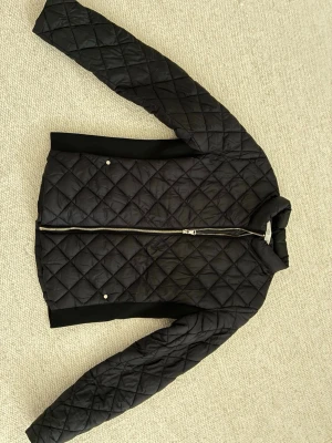 Svart quiltad jacka med dragkedja - Snygg svart quiltad jacka med dragkedja framtill och två fickor med tryckknappar. Jackan har ståkrage och ribbade muddar vid ärmslut och nederkant. Perfekt för kyliga dagar och enkel att matcha med olika outfits.
