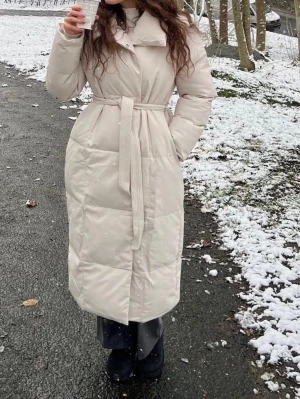 Lång beige dunjacka med bälte - Snygg lång beige dunjacka med bälte i midjan och hög krage. Jackan har quiltad design, långa ärmar och knäppning framtill. Perfekt för kalla vinterdagar och ger en clean och stilren look. Köpt på Chelsea i Göteborg. Liten fläck syns på sista bilden