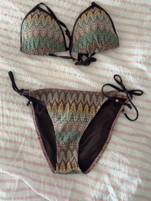 Mönstrad bikini med knyt i sidorna - Snygg bikini med färgglatt mönster i grönt, rosa, gult och brunt. Överdelen har trekantsform och knyt i nacken och ryggen. Underdelen har knyt i sidorna och samma mönster. Perfekt för stranden eller poolen! Toppen är 36 och underdelen 38 