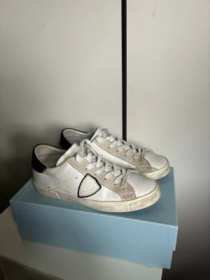 Philippe Model vita sneakers med svart detalj - Säljer ett par vita Philippe Model sneakers med svart häl och beige mockadetaljer. Skorna har en ikonisk sköldlogga på sidan, vita skosnören och en klassisk låg silhuett. Tillverkade i läder och mocka, perfekta för en clean och stilren look.