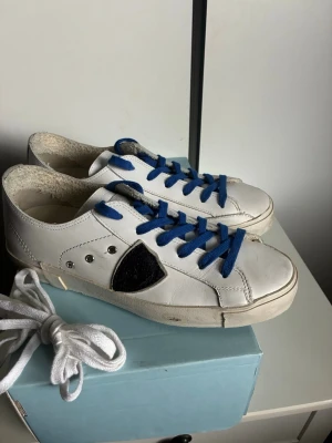 Philippe Model vita sneakers - Snygga vita sneakers från Philippe Model med blå skosnören och svart detalj på sidan. Tillverkade i läder med råa kanter runt öppningen och silverfärgade detaljer. Klassisk låg modell som passar till det mesta. Extra vita skosnören medföljer.