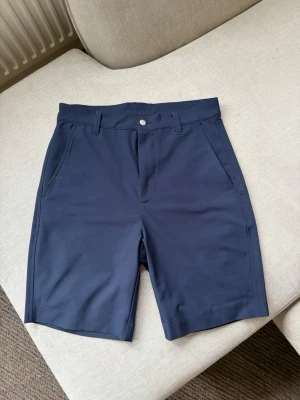 FootJoy Golfshorts – Marinblå - Säljer ett par stilrena golfshorts från FootJoy i marinblå färg. Shortsen är i storlek W30 och är i mycket fint skick. De är tillverkade i ett lätt och stretchigt funktionsmaterial som ger hög komfort och bra rörelsefrihet – perfekta för långa dagar på golfbanan.  En klassisk modell som passar lika bra till en piké på banan som till en t-shirt eller skjorta under sommaren.  Hör gärna av dig vid frågor eller om du vill ha fler bilder!