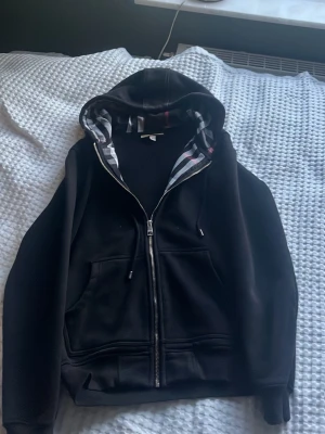 Svart hoodie med dragkedja Burberry - Säljer en svart hoodie från Burberry med dragkedja och klassiskt rutigt foder i huvan och insidan. Hoodien har fickor framtill och justerbar huva med snören. Perfekt för dig som gillar stilrena plagg med en twist. Materialet är mjukt och bekvämt.