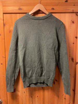 Grön stickad tröja med rund hals - En enkel och stilren grön stickad tröja med rund halsringning och ribbade muddar vid ärmslut och nederkant. Tröjan har en rak passform och är perfekt för lager-på-lager. Materialet ser ut att vara mjukt och bekvämt, troligen bomull eller ullblandning.