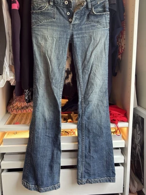 Blå bootcut jeans med knappar - Säljer ett par blå bootcut jeans med slitningar och dekorativa sömmar vid fickorna. Jeansen har knappgylf och klassisk femficksmodell. Materialet är denim i bomull med en tvättad look och ljusa detaljer. Storlek 27 men är sj det så skulle säga 26-25 eftersom de inte passar