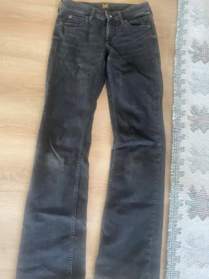 Svarta bootcut jeans från Lee - Säljer ett par svarta bootcut jeans från Lee med klassisk femficksdesign och knappgylf. Jeansen har en lätt tvättad look och raka ben som går ut något nedtill. Perfekta för dig som gillar en avslappnad men ändå snygg stil. w 26 L33
