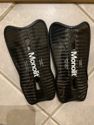 Monolit benskydd i kolfiber - Säljer ett par svarta benskydd från Monolit i kolfiber-look. De har en lätt, smidig form och två öppningar för bättre ventilation. Perfekta för dig som vill ha skydd med låg vikt och snygg design till träning eller match.