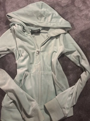 Ljusgrön velourhoodie Juicy Couture - Snygg ljusgrön hoodie i mjuk velour från Juicy Couture. Dragkedja framtill, stor huva och fickor på sidorna. Ikonisk JC-logga broderad på bröstet. Perfekt för en chill och trendig look.