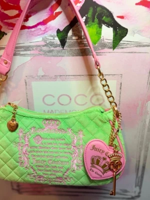 Grön och rosa Juicy Couture handväska  - Trendig quiltad handväska från Juicy Couture i limegrönt tyg med rosa detaljer och broderi. Väskan har en axelrem i rosa konstläder, guldfärgad kedja och dekorativa hängen, inklusive ett hjärta och en nyckel. Perfekt för dig som gillar färg och statement-accessoarer.
