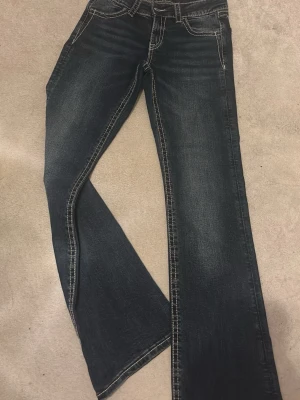 Mörkblå bootcut jeans med brodyr - Säljer ett par mörkblå bootcut jeans med snygga slitningar och kontrastsömmar. Bakfickorna har cool broderad design i beige. Klassisk femficksmodell och låg midja. Perfekta för dig som gillar en avslappnad men trendig look.