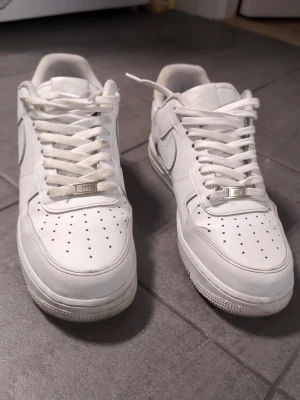 Vita Nike Air Force 1 sneakers - Klassiska vita Nike Air Force 1 sneakers med lågt skaft. Skorna har perforerad tå, vit sula och ikonisk Swoosh på sidan. Tillverkade i läder med vita skosnören och metallbricka med 'AF1' på snörningen. Perfekta för en clean och stilren look. St 42 använt ca 6 månader men fortfarande i bra skick  ordinarie pris 1299 beror på var man köper /beställer men oftast 1200 uppåt .säljes för 500