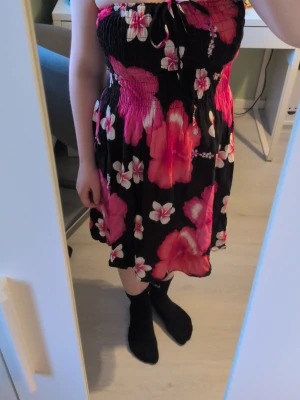 Svart och rosa blommig kortklänning - Säljer en söt axelbandslös kortklänning med svart botten och stora rosa samt vita blommor. Klänningen har smock upptill och en liten rosett vid bysten. Perfekt för varma dagar och ger en somrig vibe. Det står ingen storlek men den passar xs-m eftersom att den är så stretchig. Den går också att använda som kjol.☺️