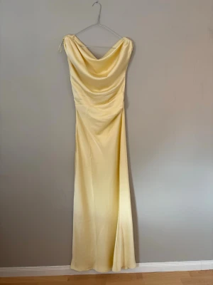 Ljusgul långklänning i satin - Elegant ljusgul långklänning i satin med draperad urringning och ärmlös design. Klänningen har en slim passform som framhäver siluetten och ett mjukt, glansigt tyg som ger en lyxig känsla. Perfekt för speciella tillfällen när du vill sticka ut.