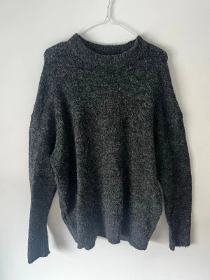 Mörkgrå stickad tröja med rund hals - Mysig mörkgrå stickad tröja med rund hals och långa ärmar. Tröjan har en avslappnad passform och är tillverkad i ett mjukt, melerat garn