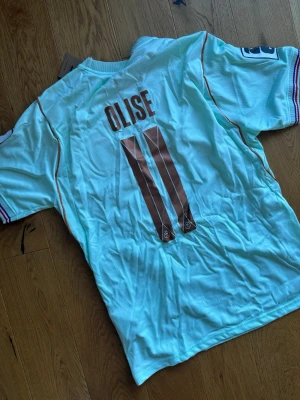 Frankrike fotbollströja Olise #11 - Ljusblå fotbollströja från Nike med Frankrikes landslagsmärke och nummer 11 Olise på ryggen. Tröjan har korta ärmar, bronsfärgade detaljer och broderad tupp på bröstet. Perfekt för dig som älskar fotboll och vill stötta Frankrike. Helt oanvänd 