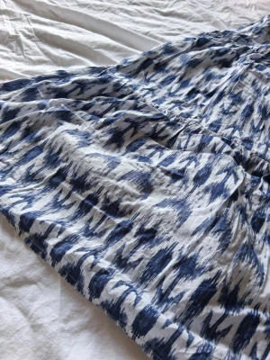 Blåvit mönstrad långkjol - Säljer en flowy långkjol i blått och vitt med ett abstrakt mönster. Kjolen har bred, smockad midja som ger en bekväm passform och är gjord i ett lätt material som passar perfekt till varma dagar.