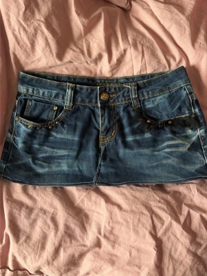 Blå jeanskortkjol med stjärndetalj - Snygg blå jeanskortkjol med slitningar och dekorativa nitar på fickorna. Kjolen har en cool stjärndetalj på ena bakfickan och klassiska jeanssömmar. Perfekt för dig som gillar en trendig och avslappnad stil.
