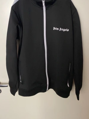 Svart zip hoodie från Palm Angels - Säljer en svart hoodie från Palm Angels med vit dragkedja och vit logga på bröstet. Hoodien har långa ärmar med vita ränder på sidan och två fickor med dragkedja. Tillverkad i ett mjukt och bekvämt material, perfekt för en avslappnad streetwear-look.
