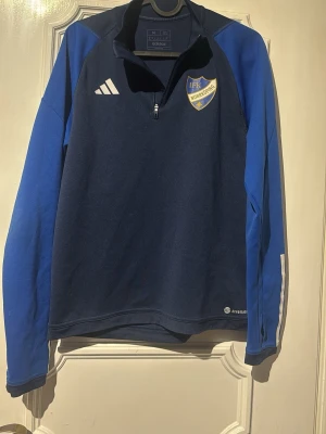 Blå IFK Norrköping träningströja Adidas - Mörkblå och blå långärmad träningströja från Adidas med IFK Norrköping-logga på bröstet och texten 'PEKING' på ryggen. Tröjan har halv dragkedja och är tillverkad i funktionsmaterial, perfekt för träning eller uppvärmning.